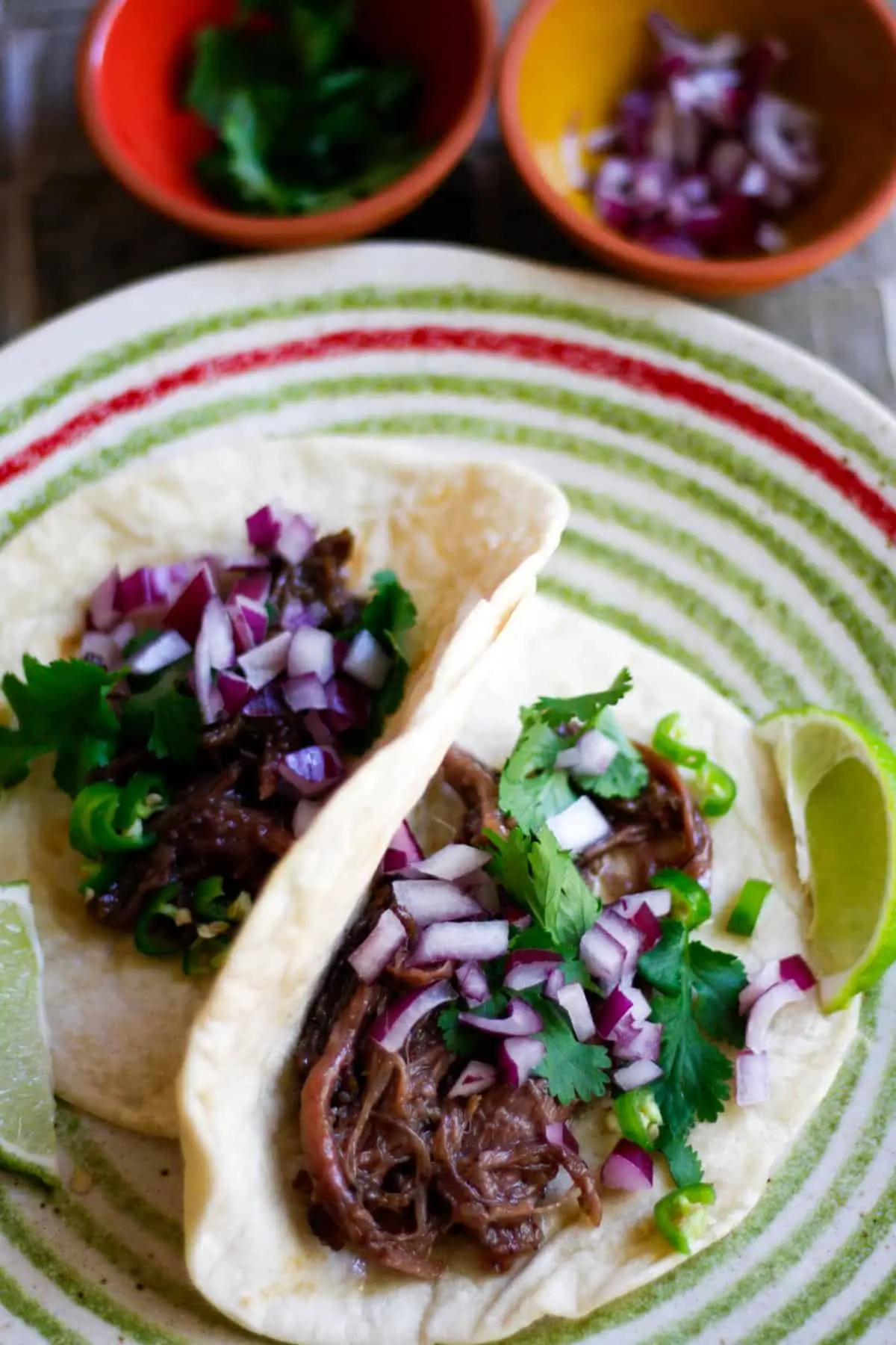 Barbacoa tacos garnished with cilantro, red onion, chilis, and lime on a plate with green and red concentric circles. There are bowls containing cilantro and diced red onion in the background.