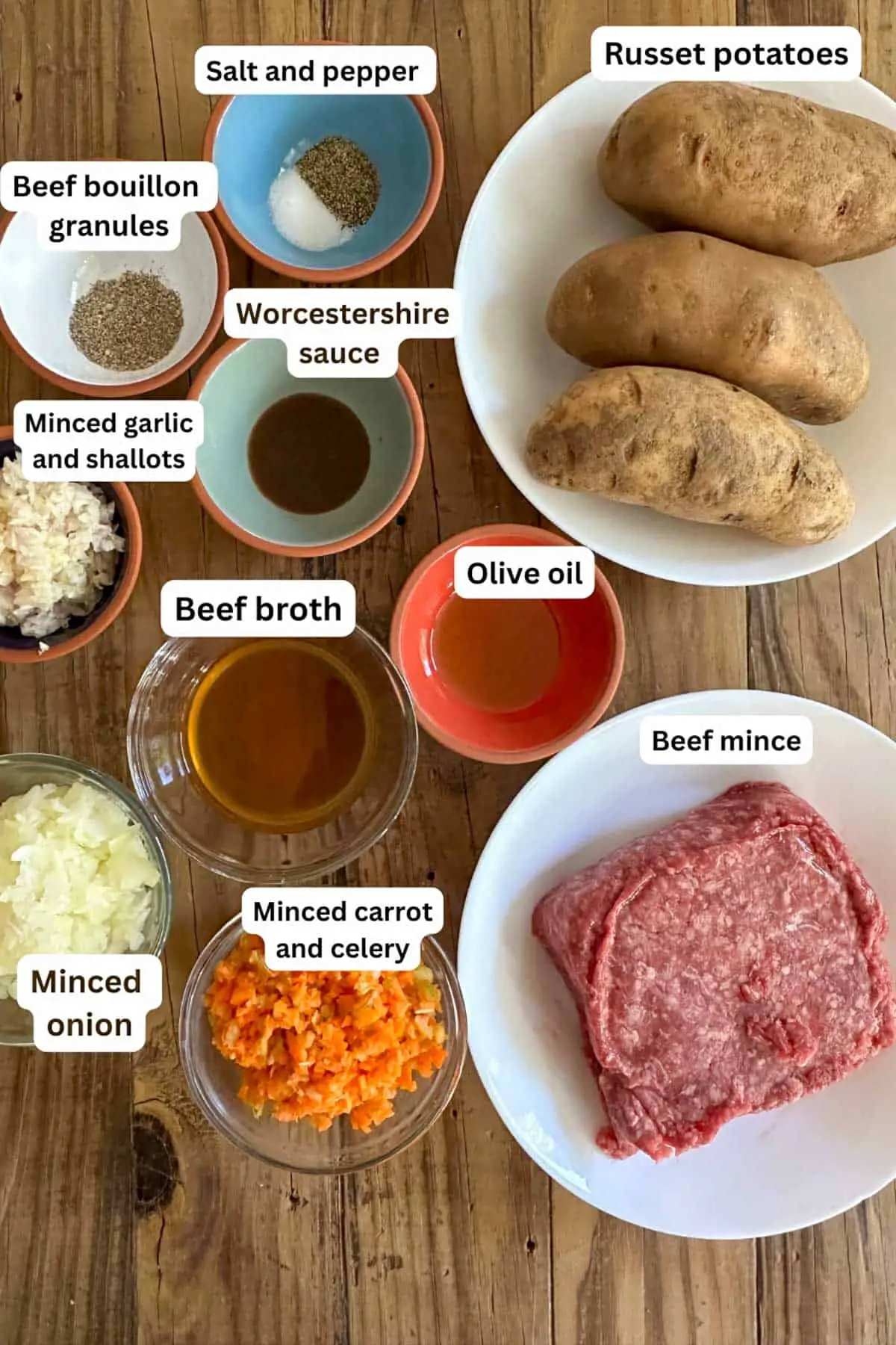 Bowls containing Russet potatoes, beef mince, olive oil, minced carrot and celery, minced onion, beef broth, minced garlic and shallots, Worcestershire sauce, beef bouillon, and salt and pepper.