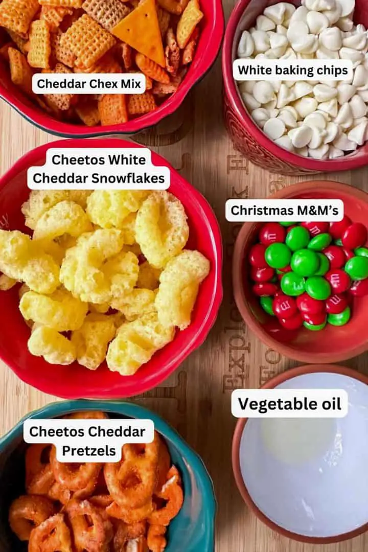 Bowls containing Cheddar Chex Mix, white chocolate baking chips, Snowflake Cheetos, red and green M&M's, Cheetos pretzels, and vegetable oil.