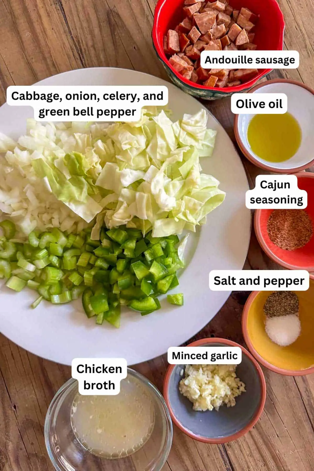 Ingredients for smothered cabbage including bowls containing Andouille Sausage, olive oil, Cajun seasoning, salt and pepper, minced garlic, chicken broth, and a plate with chopped cabbage, onion, celery and green bell pepper.