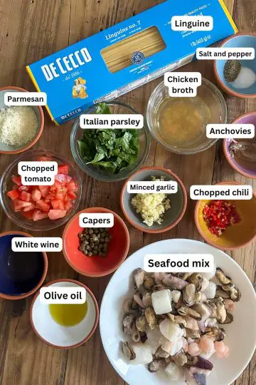 Ingredients for linguine with seafood mix including a box of linguine, and bowls containing salt and pepper, chicken broth, anchovies, Italian parsley, Parmesan, chopped tomato, white wine, capers, garlic, chili, olive oil, and seafood mix.
