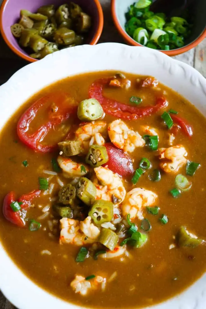 A white bowl containing shrimp gumbo with stewed tomatoes and sliced okra and green onions as garnish. There are small bowls with sliced okra and sliced green onions at the top of the bowl.