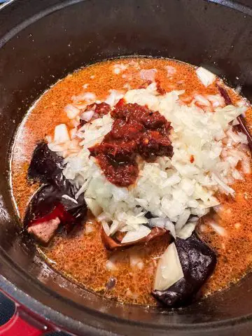 A large pot containing posole, pork seasoned broth, minced onion and garlic, red chile peppers, and red chile sauce.