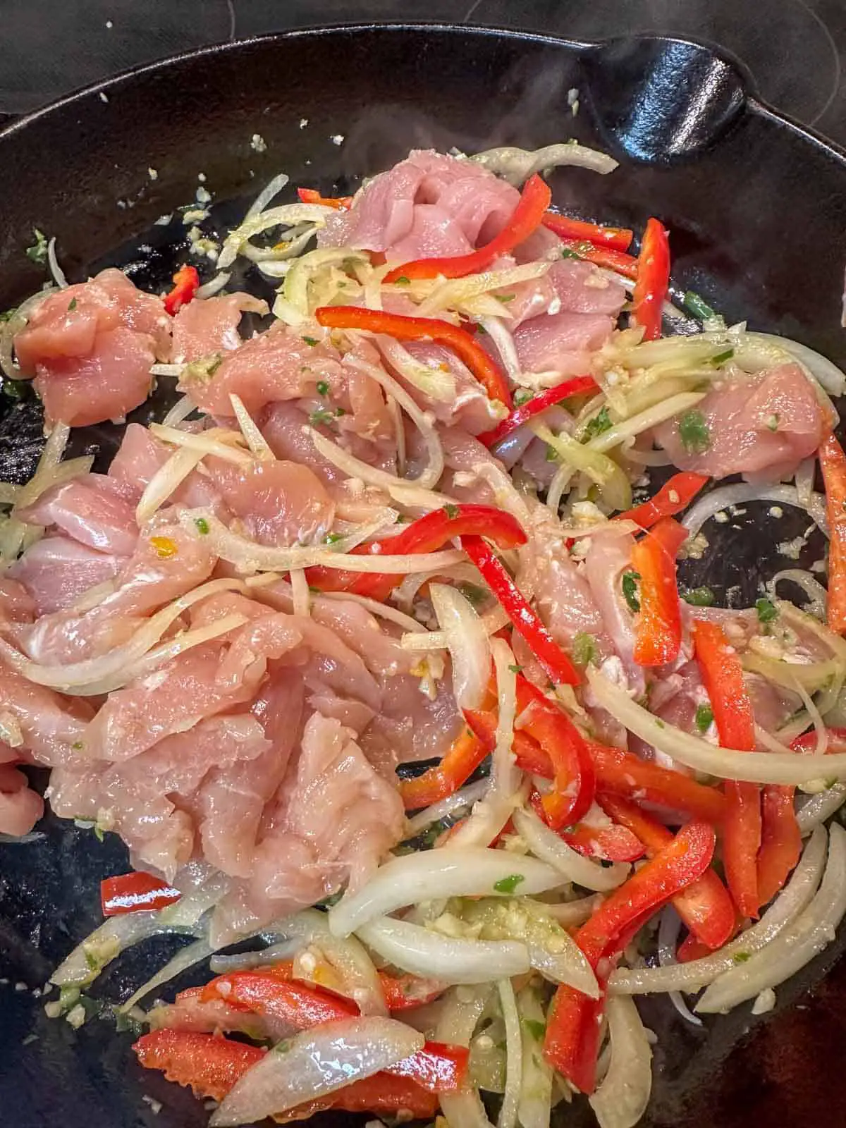 Raw sliced chicken, sliced red bell pepper, sliced onion, and minced garlic and chilis in a cast iron pan.