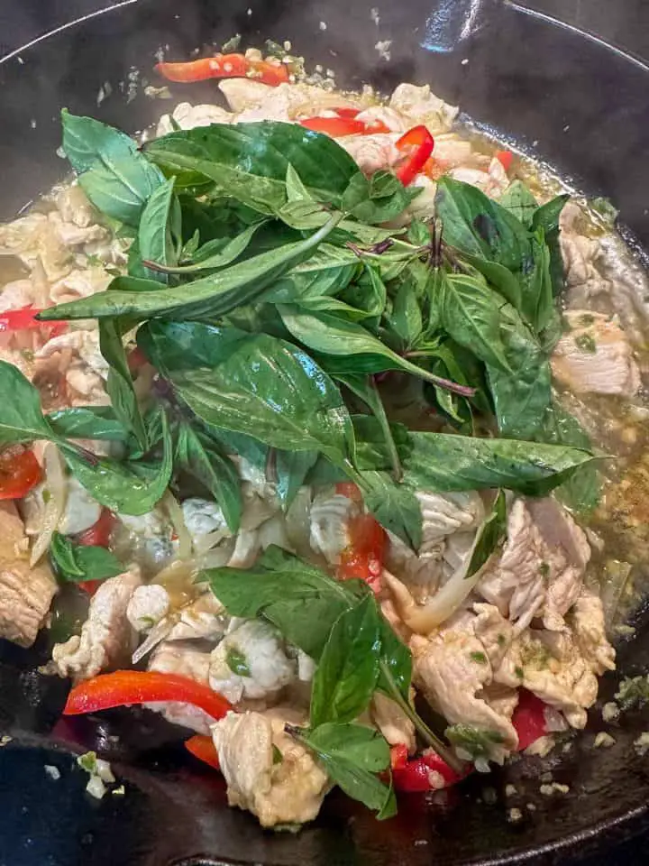 A cast iron pan containing sliced chicken, red bell pepper, onions, and minced garlic and chilis, topped with Thai basil.