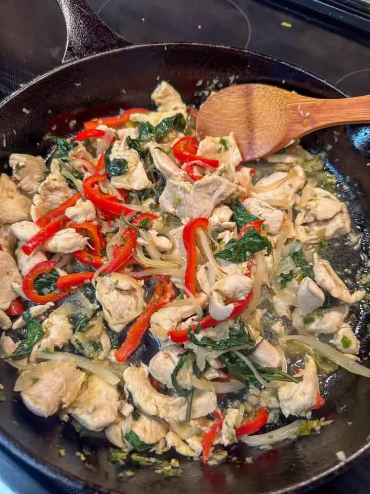 Sliced chicken, red bell pepper, onions, and Thai basil in a cast iron pan with a wooden spoon.