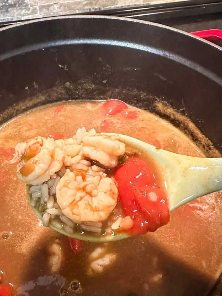A large pot containing shrimp gumbo. A blue spoon containing some of the gumbo is poised over the broth. The spoon contains shrimp, rice, and stewed tomatoes.