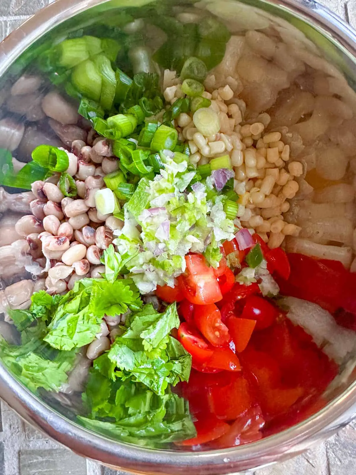 A metal bowl containing shoepeg corn, black eyed peas, green onions, cilantro, grape tomatoes, and minced garlic, onion, and jalapeno pepper.
