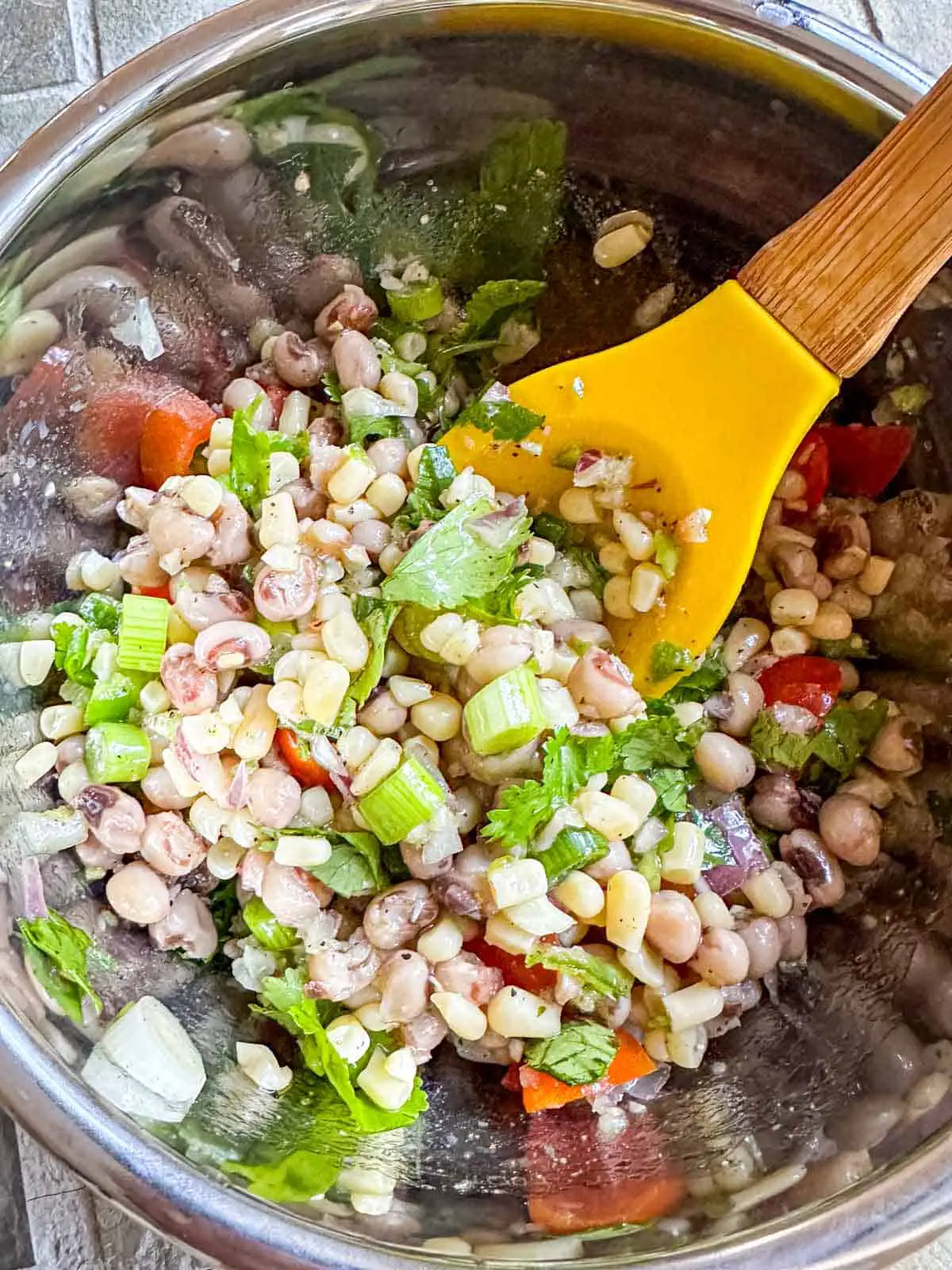 A metal bowl containing Texas caviar which is shoepeg corn, black eyed peas, green onions, cilantro, grape tomatoes, and minced garlic, onion, and jalapeno pepper. This mixture has been combined and there is a yellow spoon with a wooden handle in the bowl.