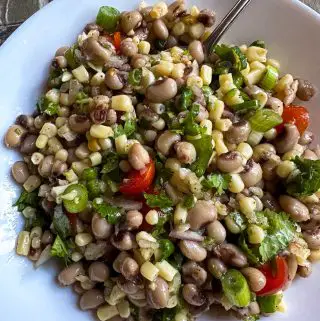 Texas caviar which is a mixture of shoepeg corn, black eyed peas, green onions, cilantro, grape tomatoes, and minced garlic, onion, and jalapeno pepper. The Texas caviar is in a white bowl and there is a spoon in the bowl.