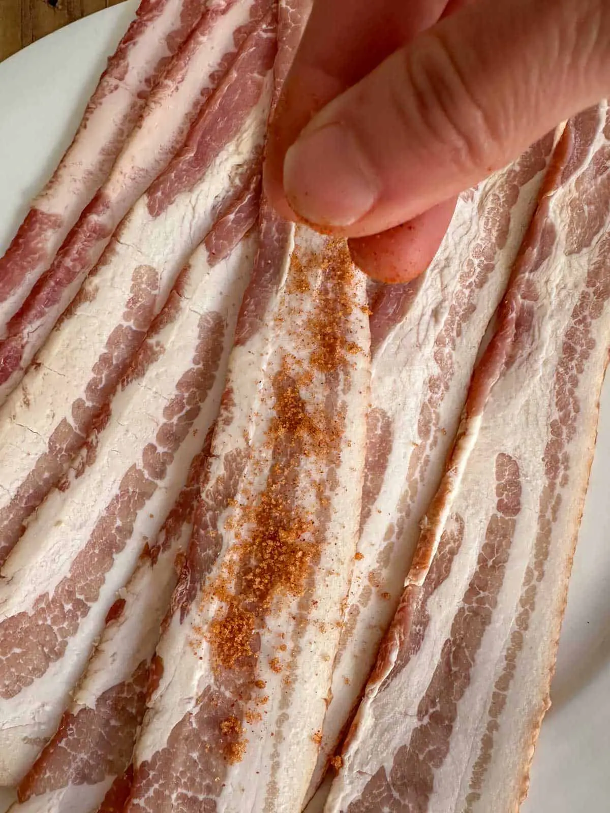Uncooked bacon on a white plate. There is a person sprinkling a mixture of Scorpion pepper powder and brown sugar onto the bacon.