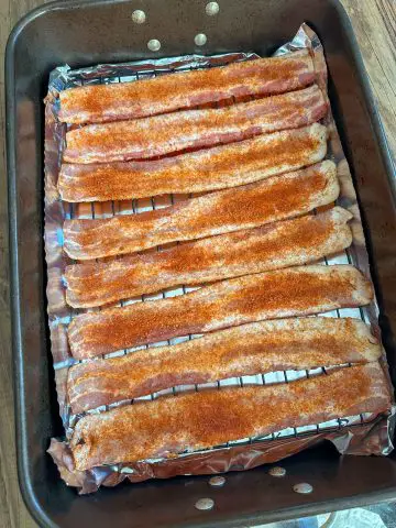 Uncooked bacon sprinkled with scorpion pepper powder and brown sugar. The bacon is on a rack over a foil lined baking pan.