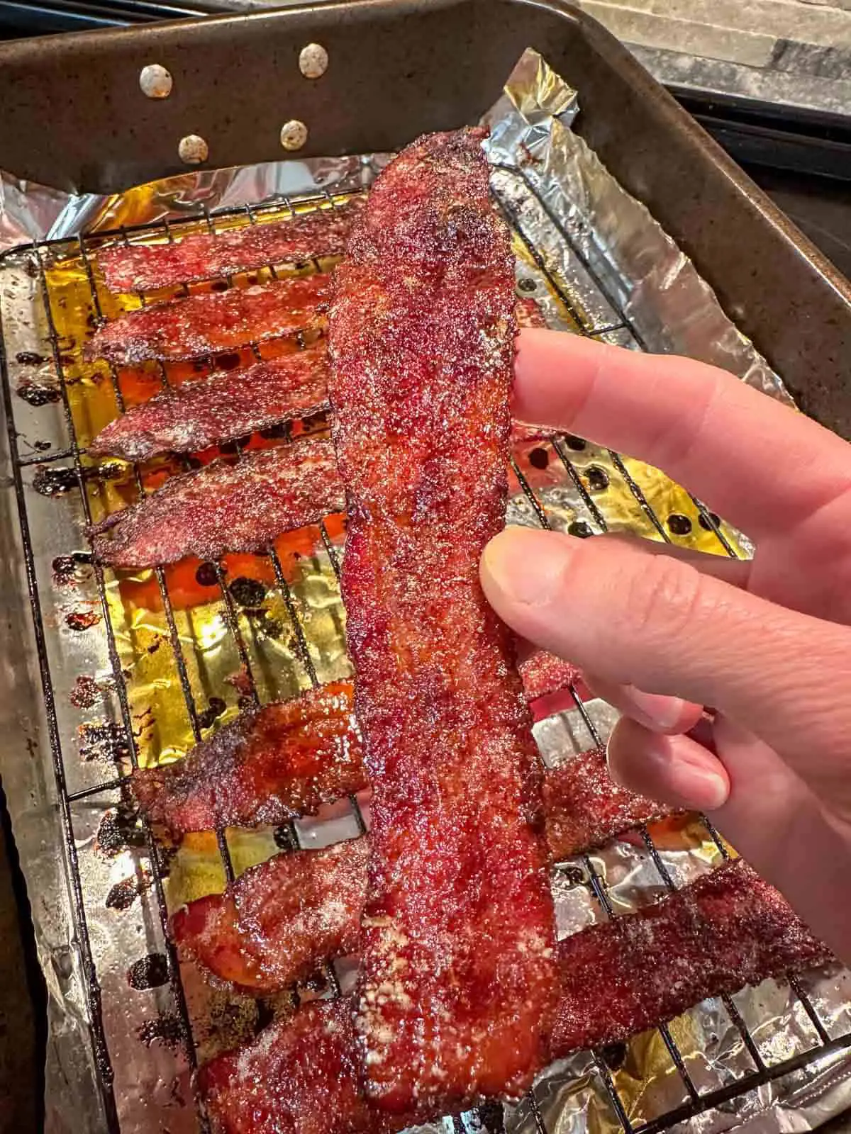 A person holding a piece of Scorpion Pepper Bacon. Behind this is a roasting tray with rack containing more Scorpion Pepper Bacon over foil with drippings underneath.