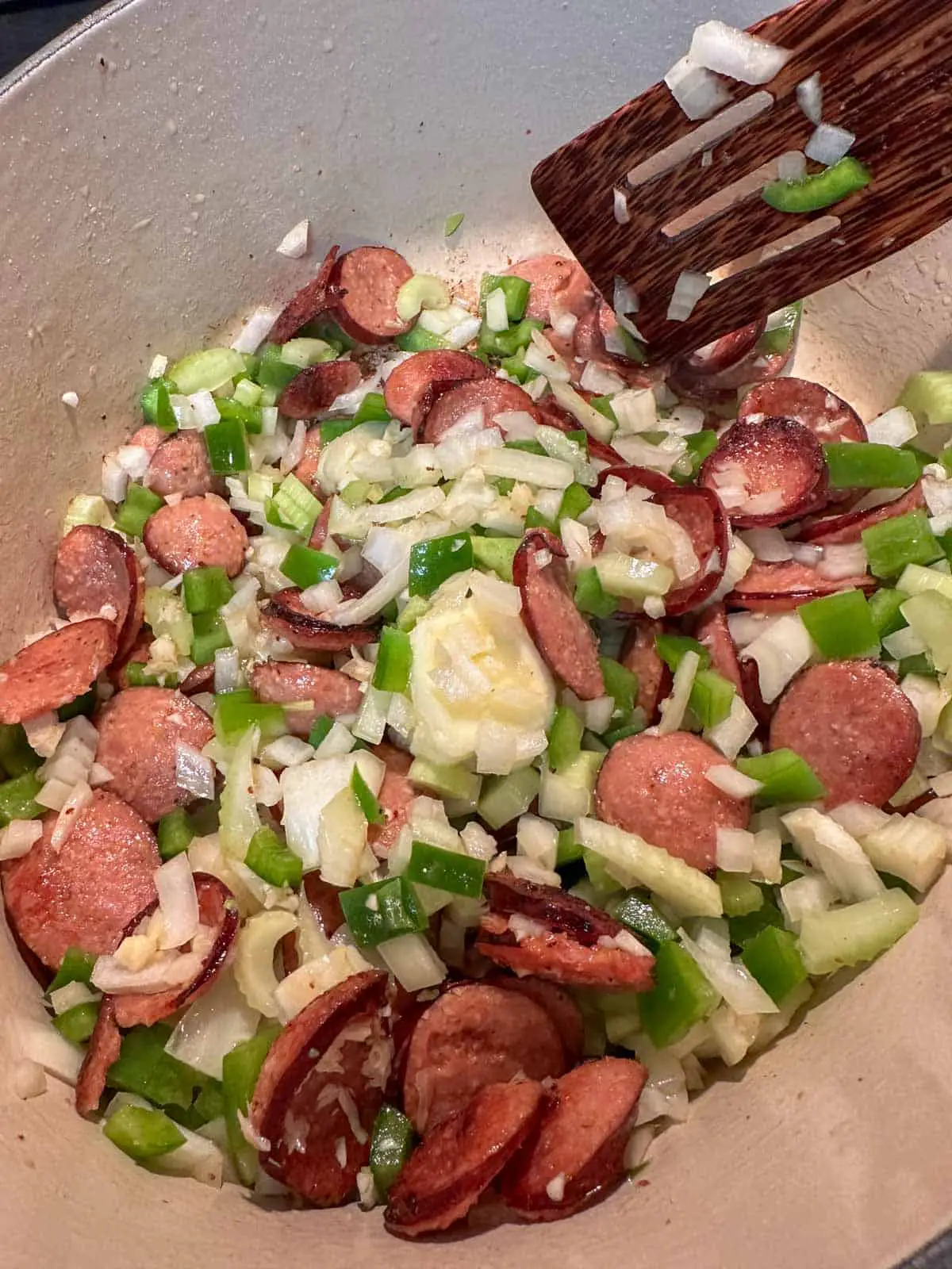 A large pot containing sliced sausage, celery, onion, and green bell pepper. There is a wooden spoon in the pot.