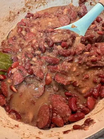 A large pot containing creamy New Orleans style red beans with sliced sausage and a bay leaf. There is a blue spoon in the pot.