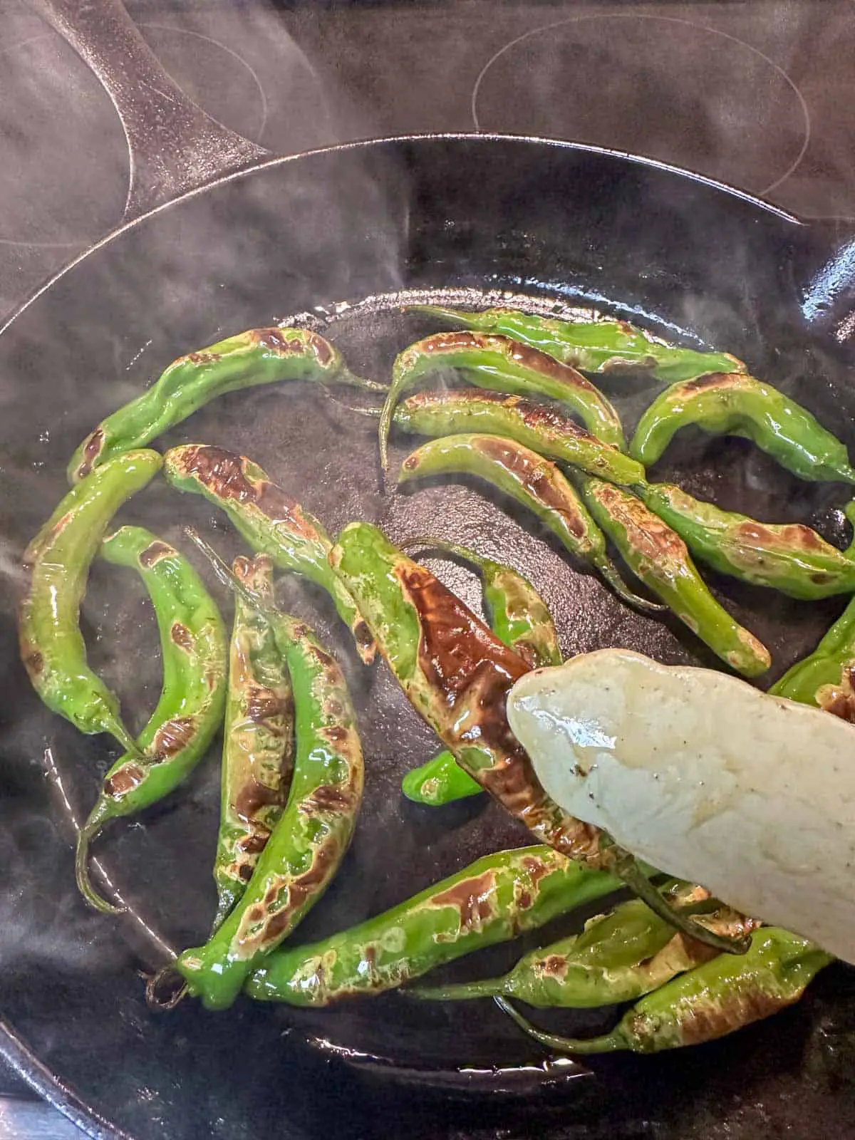 Blistered shishito peppers in a cast iron pan with smoke. There is a blue pair of tongs holding one of the shishito peppers.