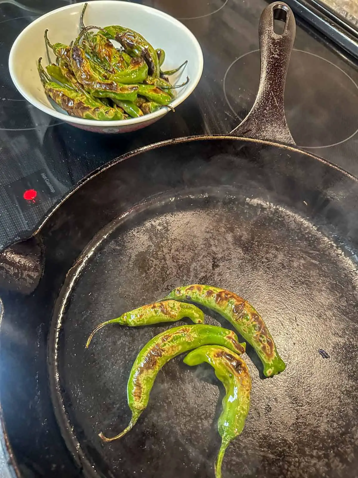 A cast iron pan containing some blistered shishito peppers on a stove. There is a small bowl containing some additional shishito peppers.