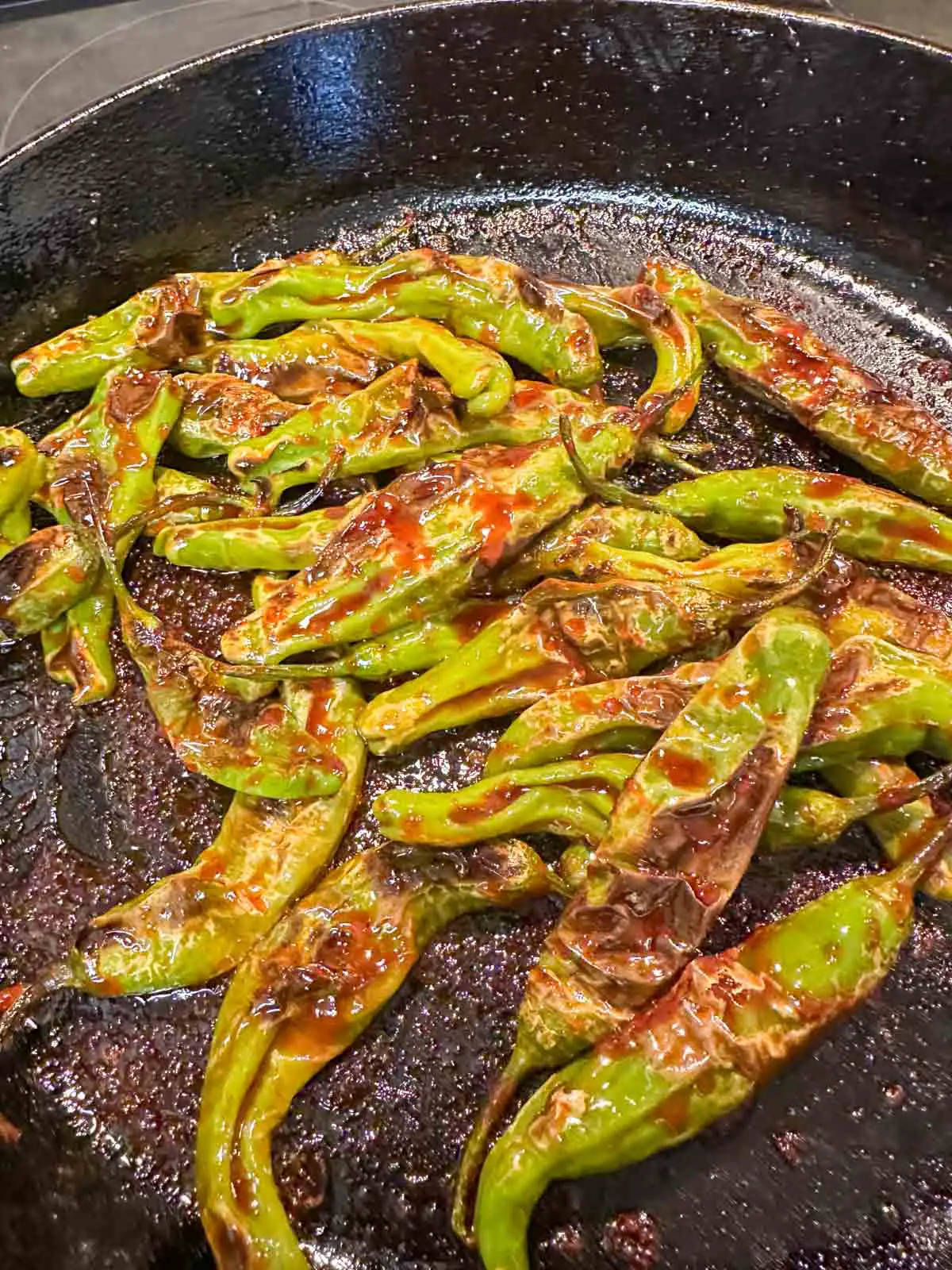 Blistered shishito peppers cooked with a spicy gochujang sauce in a cast iron pan.