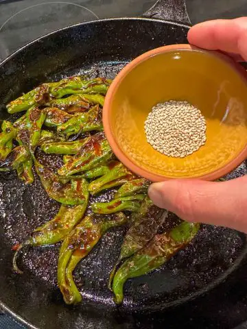 Blistered shishito peppers cooked with a spicy gochujang sauce in a cast iron pan. Theer is a person holding a small bowl containing sesame seeds above the pan.