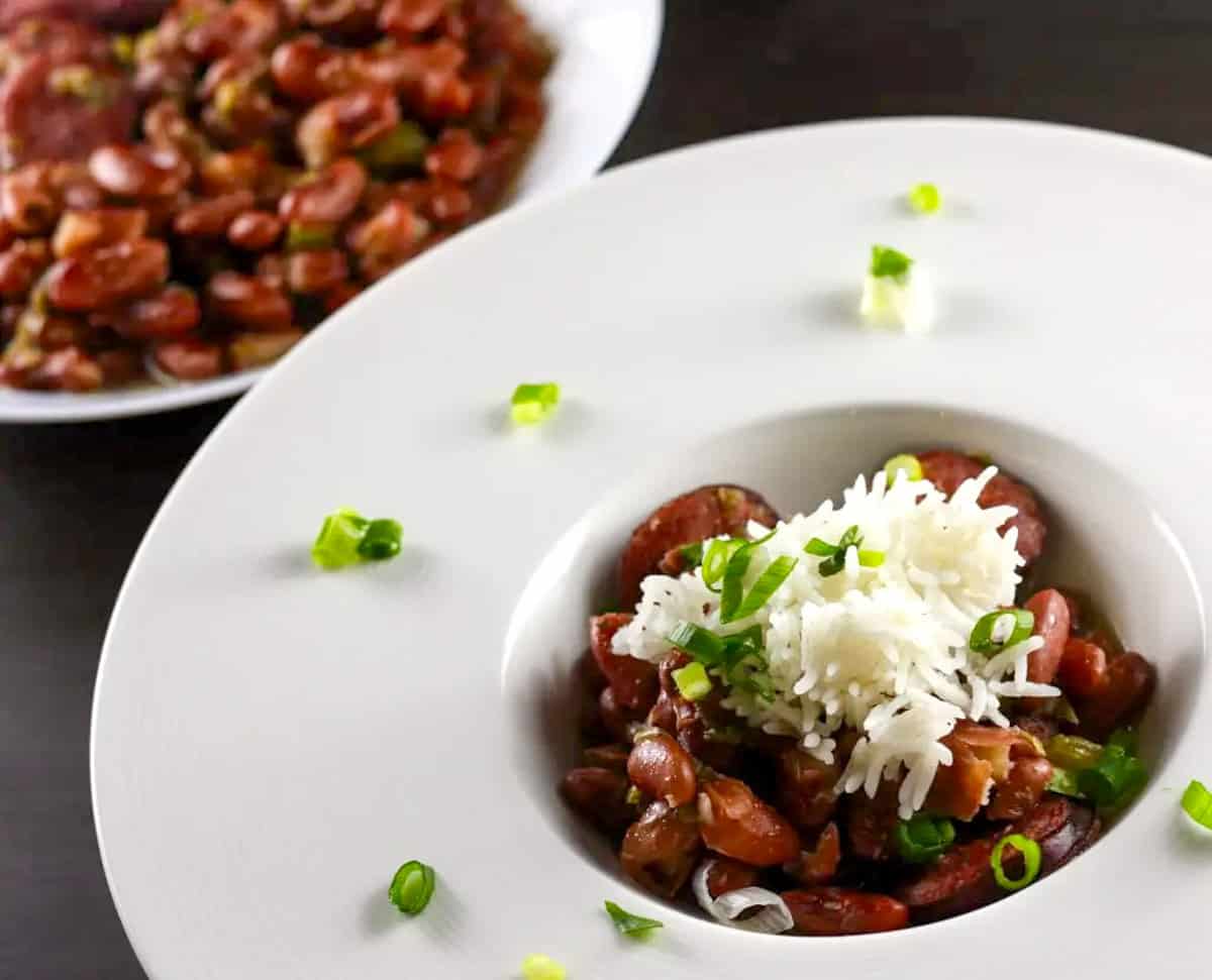 A white dish containing red beans and rice garnished with sliced green onions. There is another dish with red beans in the background on the top left.