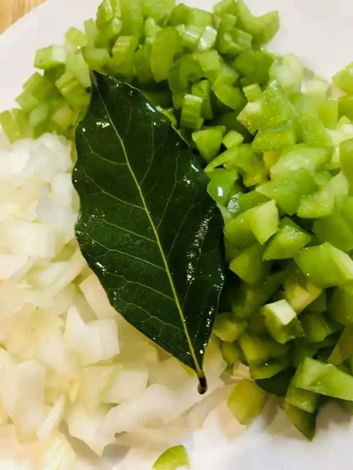 Diced onion, green bell pepper and celery and a bay leaf on a white plate.