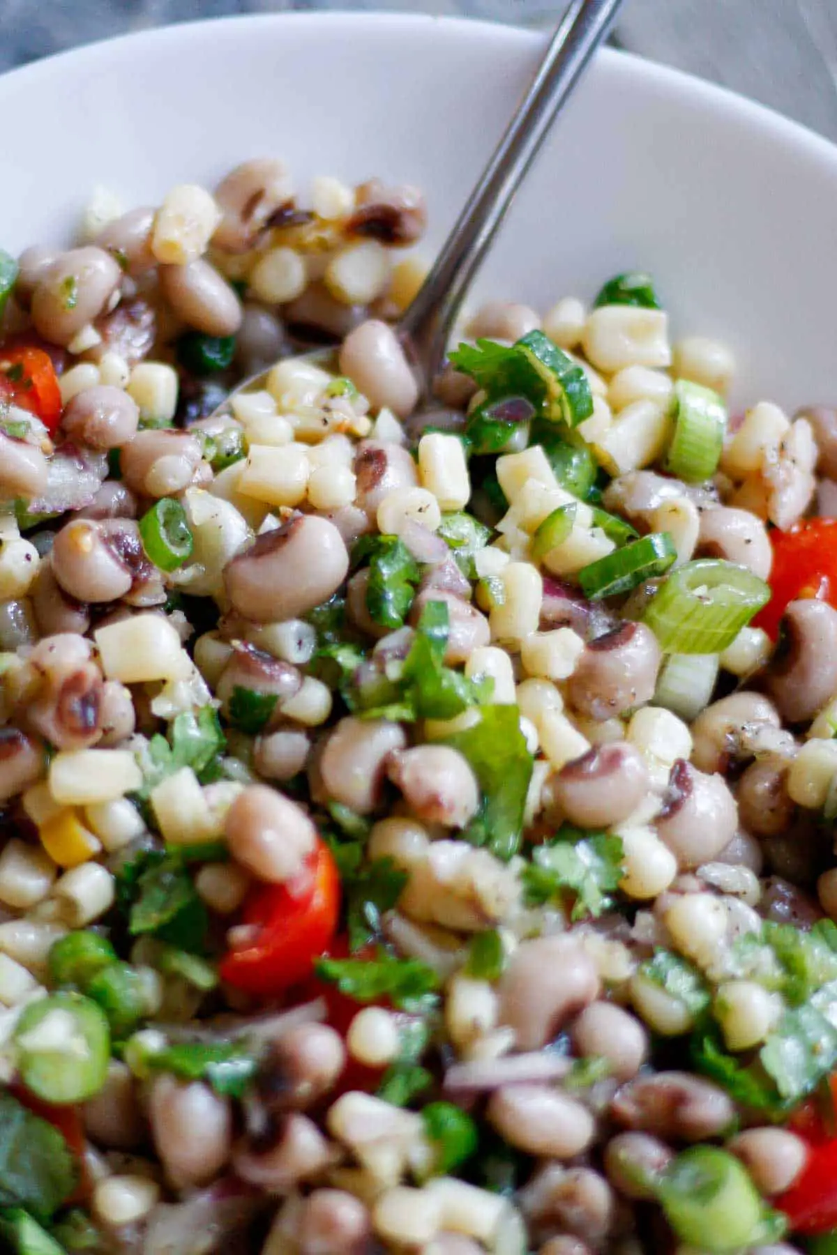 Texas caviar which is a mixture of shoepeg corn, black eyed peas, green onions, cilantro, grape tomatoes, and minced garlic, onion, and jalapeno pepper. The Texas caviar is in a white bowl and there is a spoon in the bowl.
