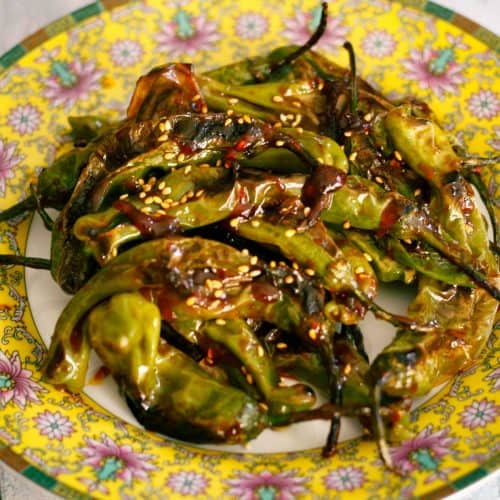 A patterned plate containing blistered shishito peppers covered with a spicy gochujang sauce garnished with sesame seeds.