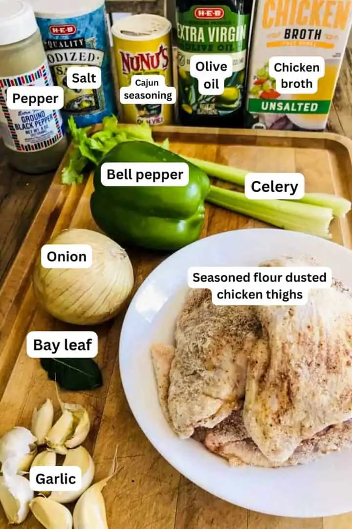 Ingredients for Cajun Smothered Chicken including seasoned flour dusted chicken thighs, garlic cloves, bay leaf, onion, bell pepper, celery stalks, chicken broth, olive oil, Cajun seasoning, salt, and pepper.