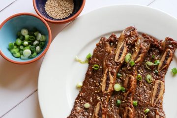 Korean Short Ribs garnished with sesame seeds and green onions on a white plate. There are small bowls on the side containing green onions and sesame seeds.