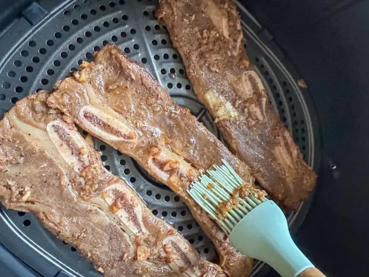 Korean beef short ribs in an air fryer basket. Someone is using a baster to baste one of the short ribs with additional marinade.