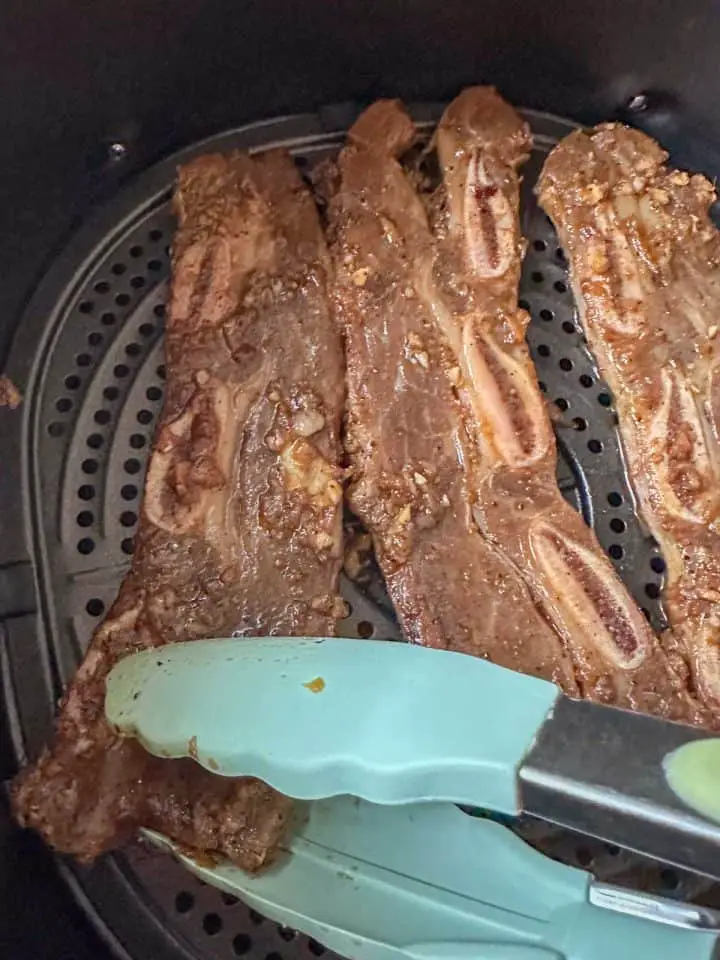 Korean beef short ribs in an air fryer basket. There is a set of tongs in the process of turning one of the beef short ribs over.