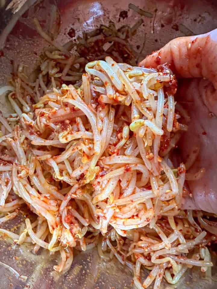 Korean spicy bean sprouts in a mixing bowl which have been mixed by hand. There is a hand holding some of the mixed bean sprouts visible in the picture.