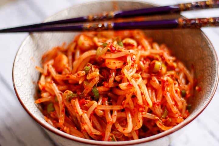 Korean Spicy Bean Sprouts in a patterned bowl with a pair of purple chopsticks resting on top of the bowl.