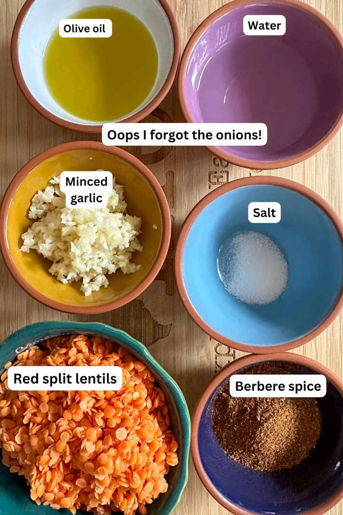 Bowls containing ingredients needed for Ethiopian Red Split Lentil Wat including olive oil, water, minced garlic, salt, red split lentils, and Berbere spice. There is text overlay stating "oops I forgot the onions!"