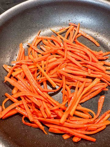 Julienned carrots being cooked in oil in a skillet.
