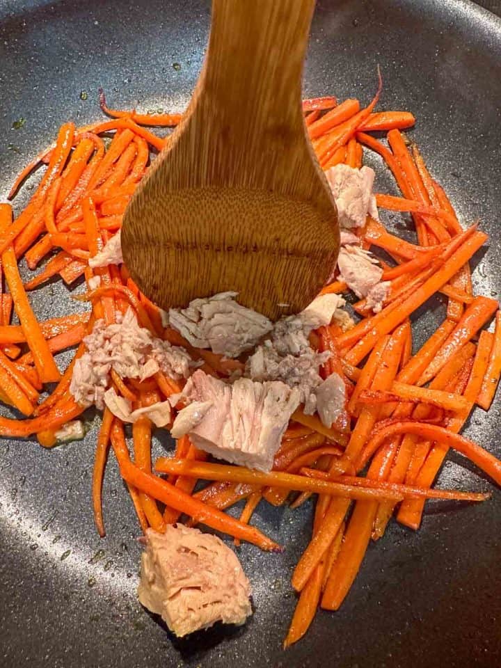 Julienned carrots and tuna in a skillet. There is a wooden spoon breaking up some of the tuna.