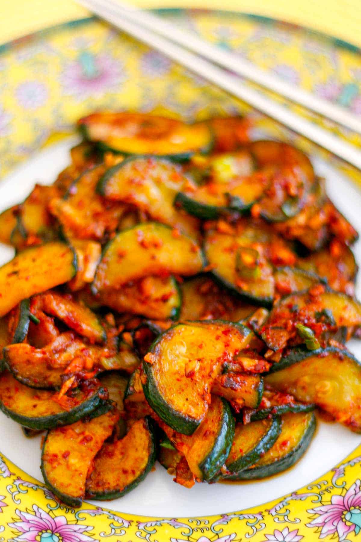 Slices of zucchini stir fried with green onions and Korean red pepper flakes place on a patterned plate with a pair of silver chopsticks resting on the right side of the plate.