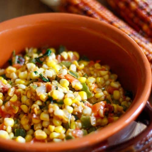 Cajun Corn Maque Choux including corn, green pepper, parsley, and tomato in a terracotta bowl. There are ears of dried corn in the background.