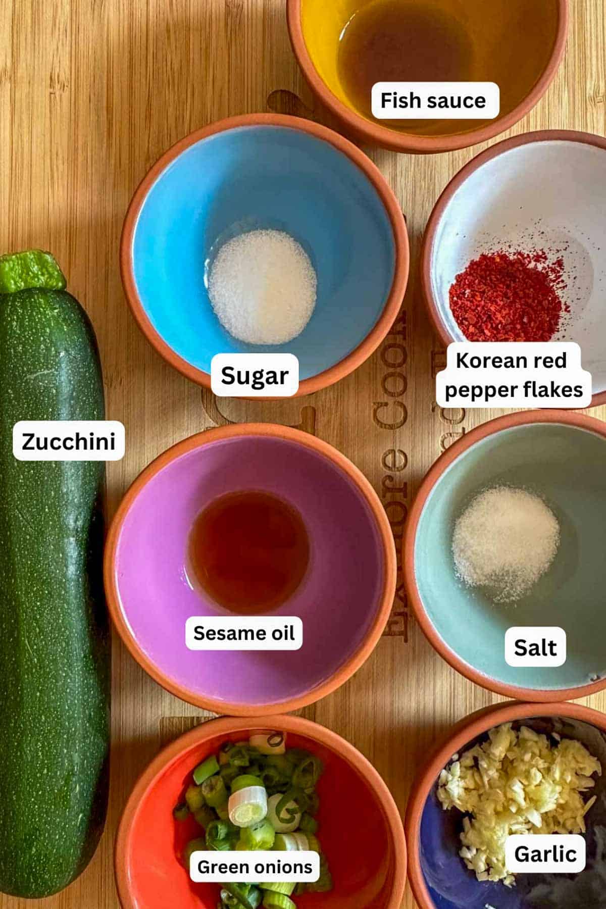Ingredients for Korean Zucchini Side Dish displayed in small bowls including fish sauce, sugar, Korean red pepper flakes, sesame oil, salt, green onions, and garlic. There is also one zucchini.