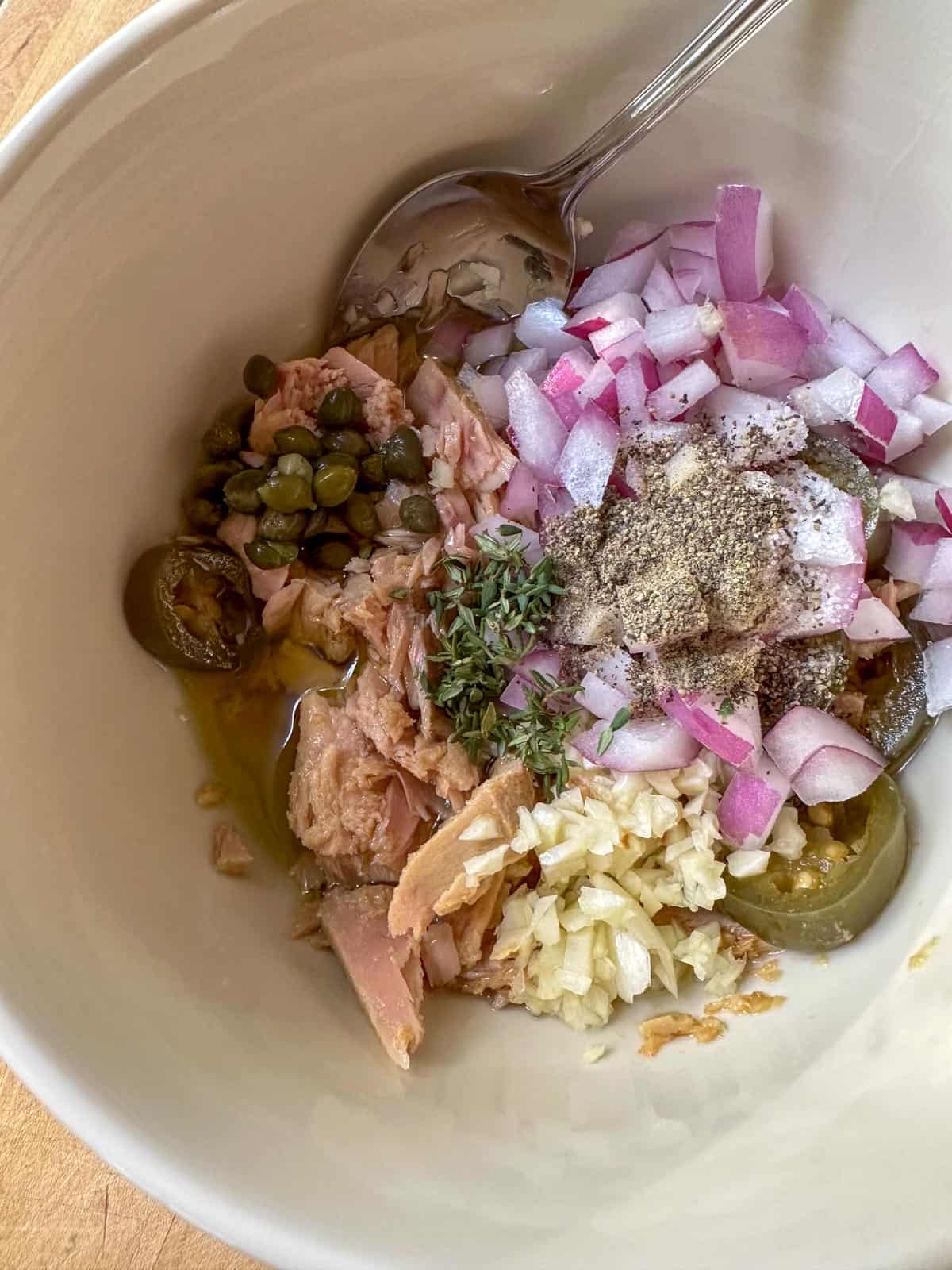 A white bowl containing diced red onion, salt and pepper, jalapeno, minced garlic, capers, tuna, and thyme. There is a spoon resting in the bowl.