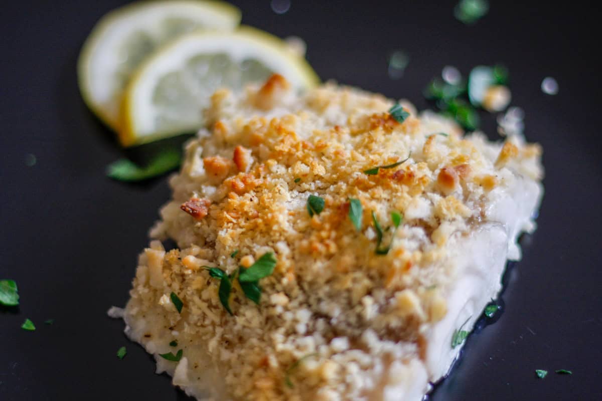 A Macadamia Nut Crusted Barramundi fillet with a golden brown crust on a black plate with lemon slices and parsley garnish.