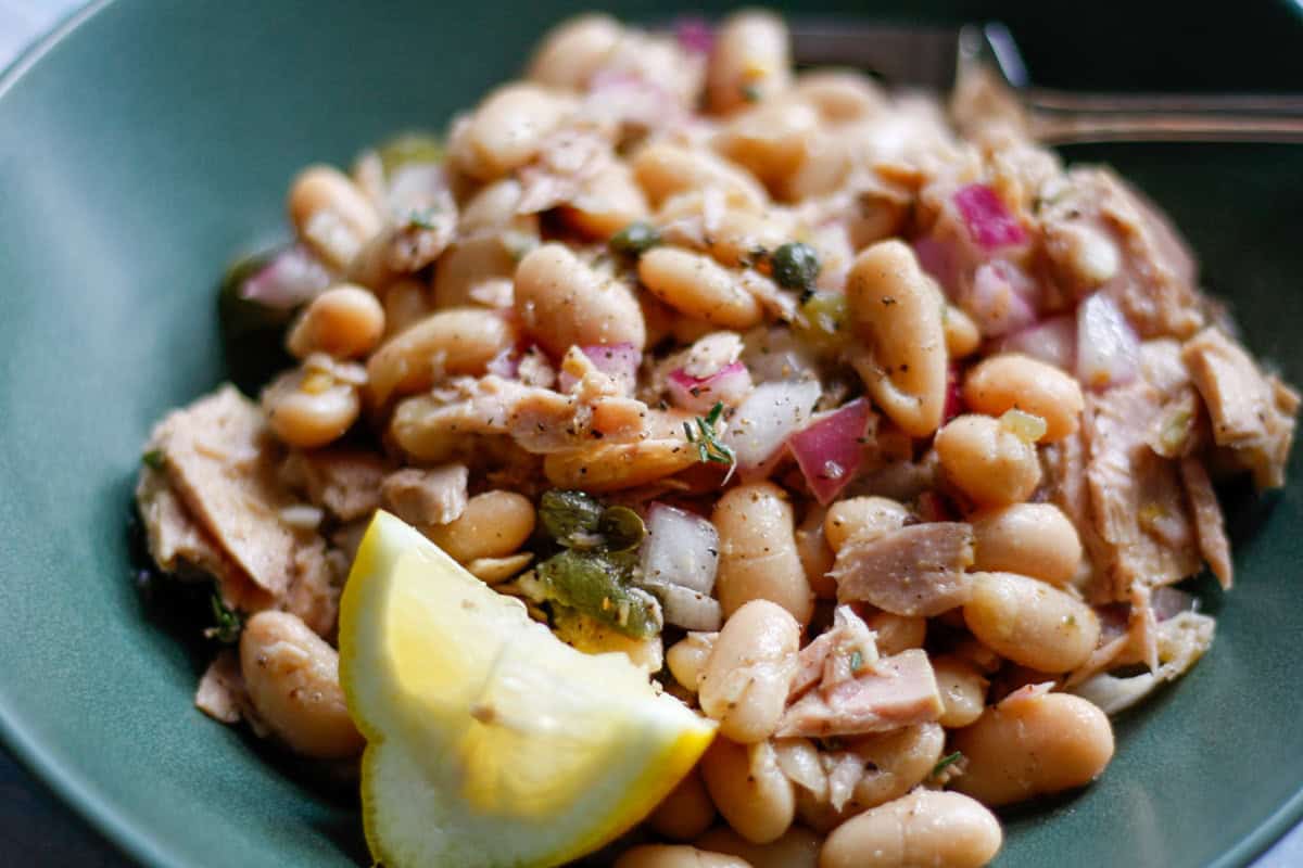 Cannellini White Bean Salad With Tuna in a green bowl. The salad also includes capers red onion, and thyme, and there is a wedge of lemon next to the salad.