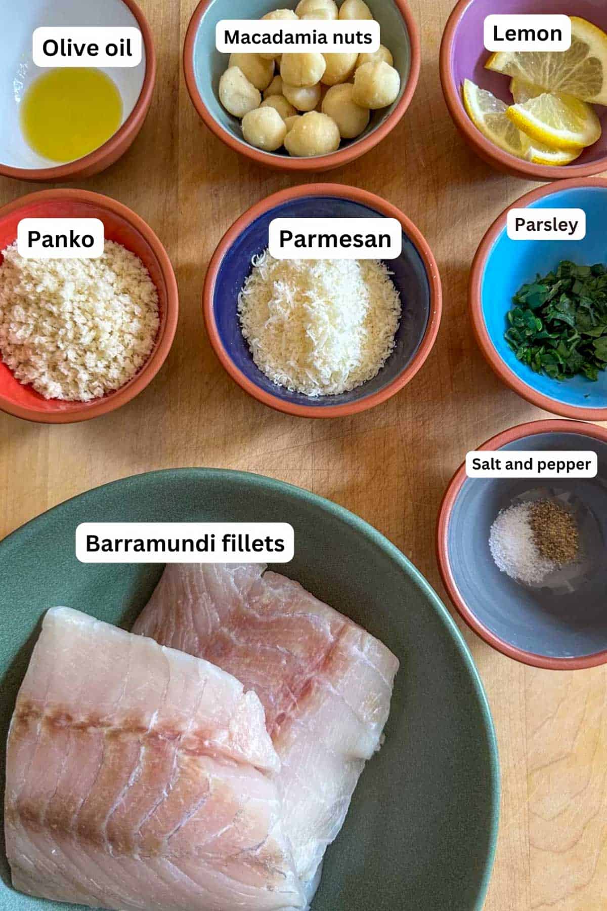 Ingredients needed to make Macadamia Nut Crusted Barramundi including olive oil, macadamia nuts, lemon slices, panko, Parmesan, parsley, barramundi fillets, and salt and pepper all displayed in bowls.