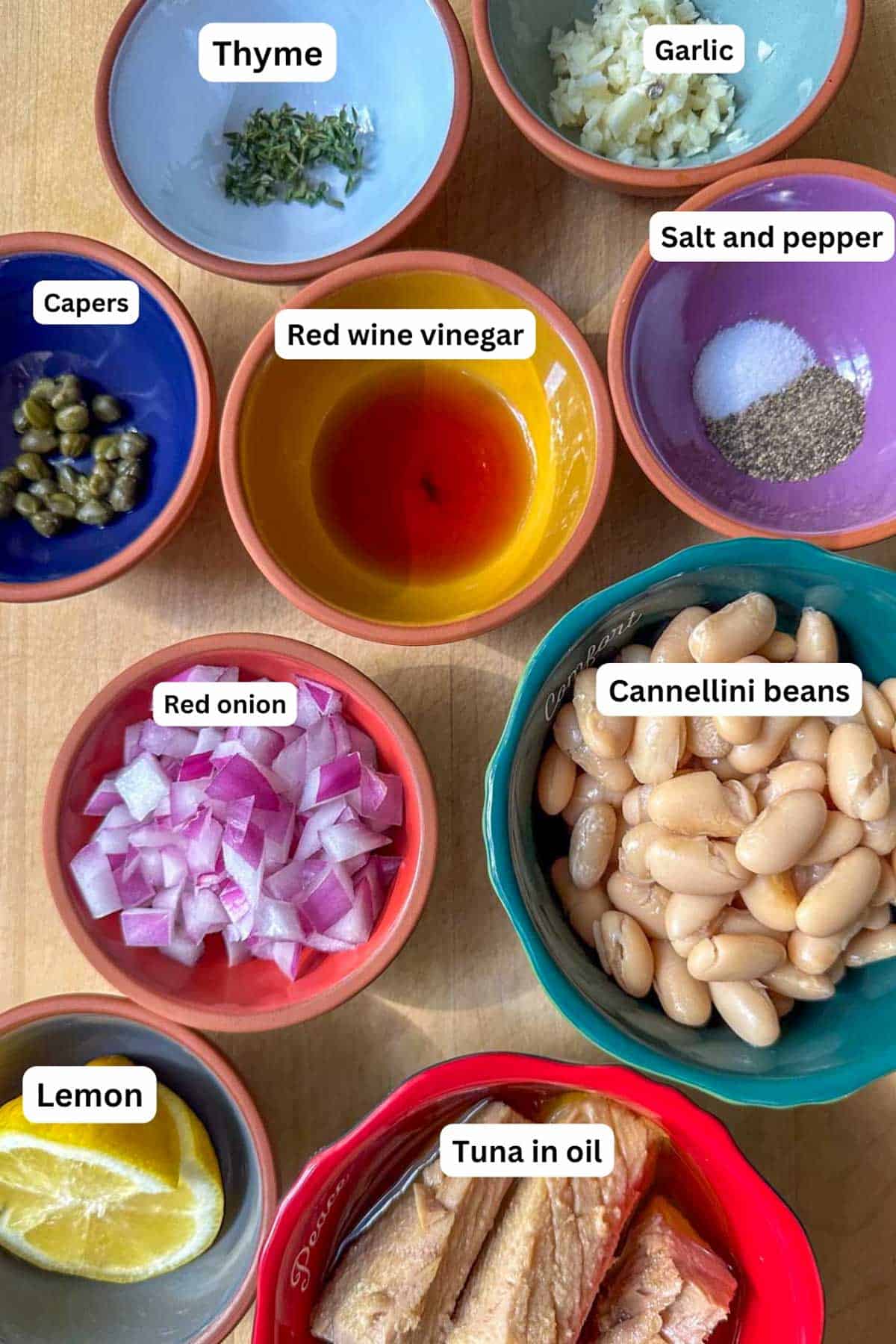 Ingredients for an Easy Cannellini White Bean Salad With Tuna Recipe displayed in bowls. They are thyme, garlic, capers, red wine vinegar, salt and pepper, red onion, cannelini beans, lemon, and tuna in oil.
