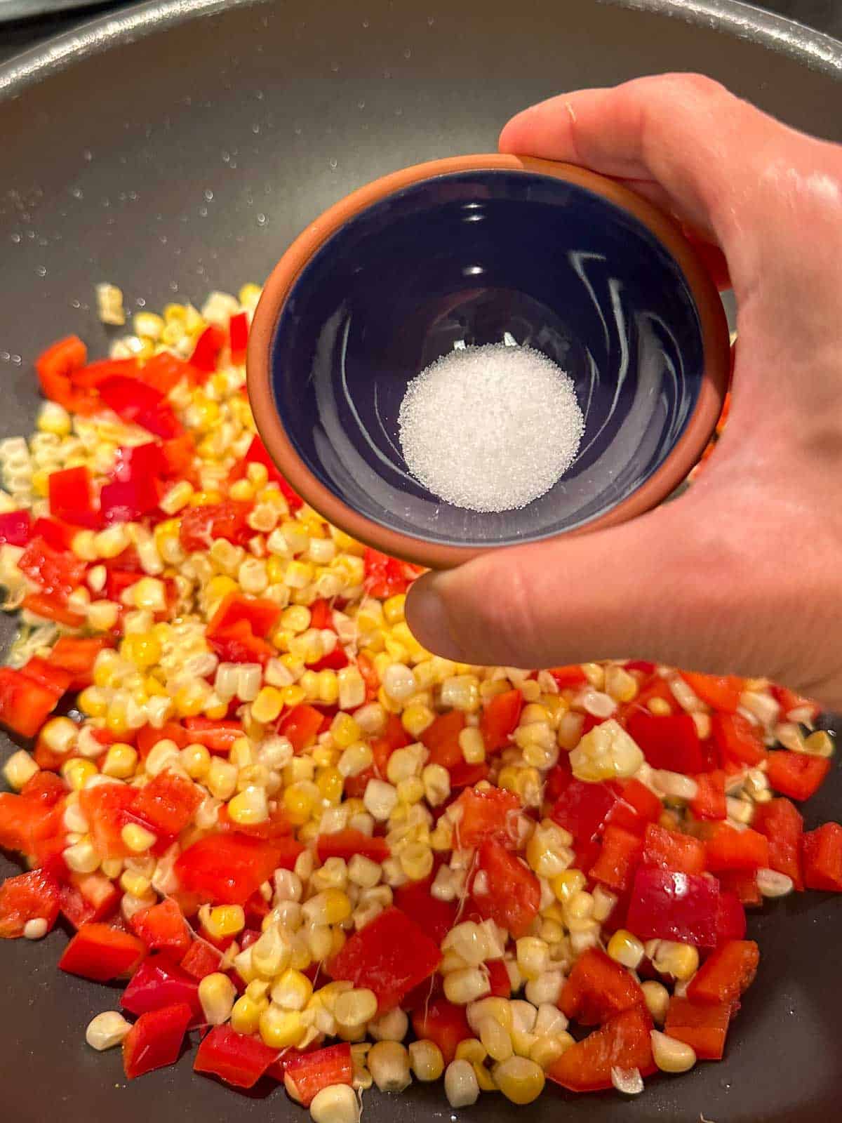 A wok containing Chinese fried corn with diced red bell peppers. There is a person holding a small bowl containing salt above the wok.