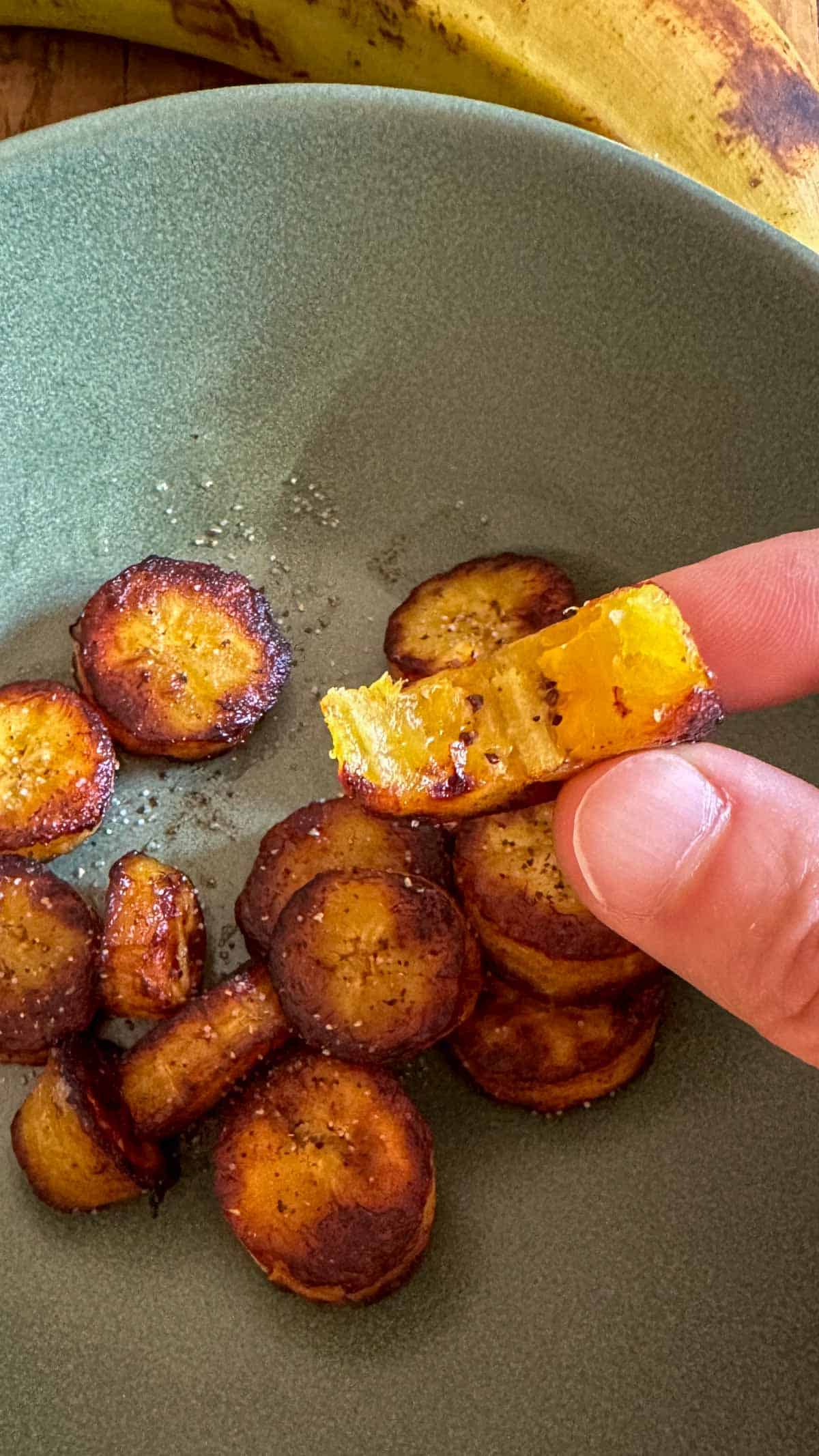 Sweet Plantains that have been sliced, fried in butter, and seasoned with salt and pepper. One of the slices has been bit into so that you can see the tender middle. This slice is in someone's hand.