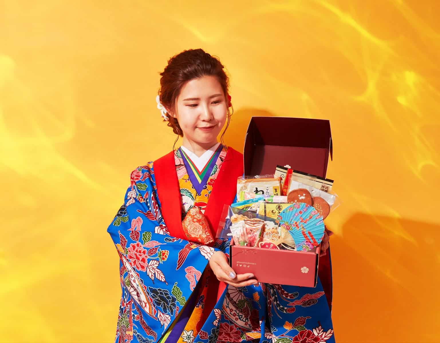 A woman dressed as a geisha holding an opened Sakuraco box showing the contents of the box.