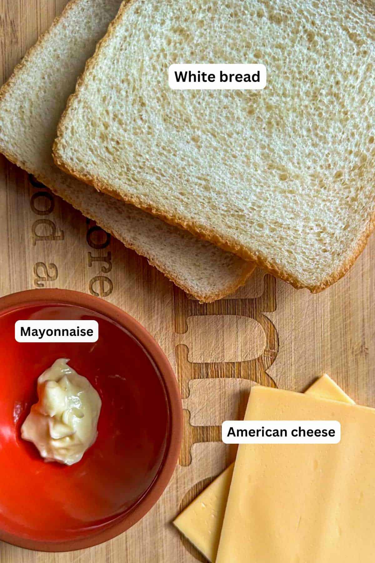 Two pieces of white bread stacked on top of each other next to a bowl with mayonnaise and two slices of Kraft American Deli Deluxe Cheese.