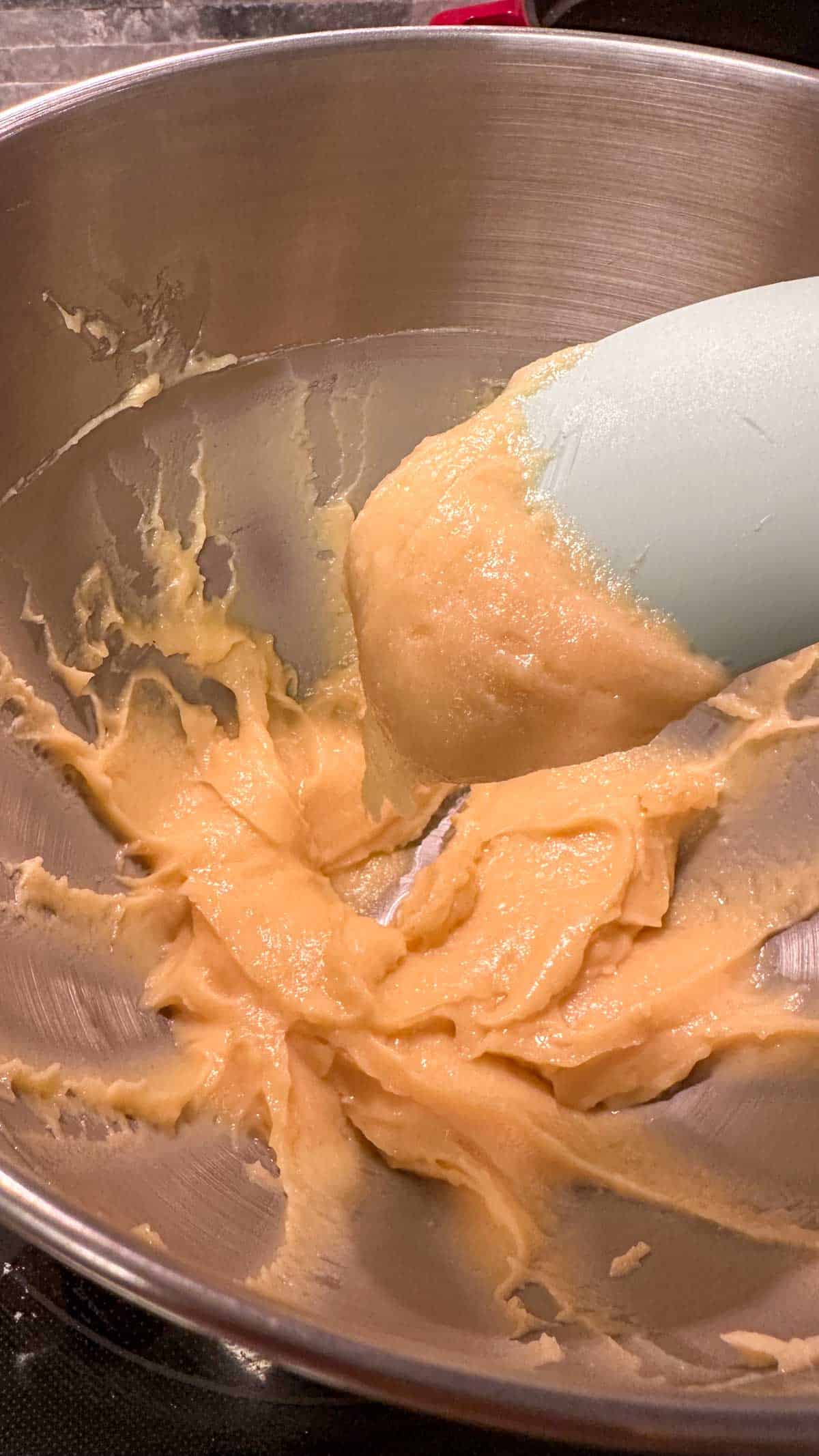 A stand mixer bowl containing the dough mixture to be used to make Parmesan cheese drop biscuits. A blue spoon with some of the dough clinging on to it is poised above the dough in the bowl.