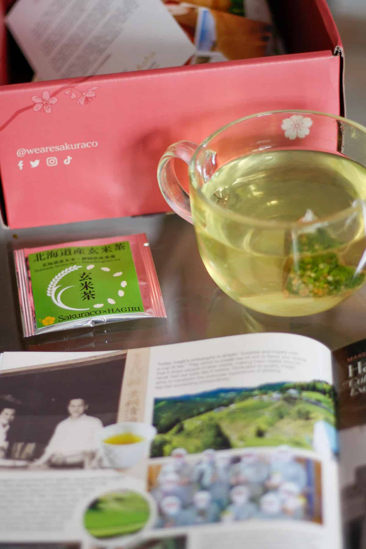 A Sakuraco box in the background with a packet of green tea and a cup of brewing green tea in front of the Sakuraco box. An opened Snack Guide from the Sakuraco box is in the foreground.